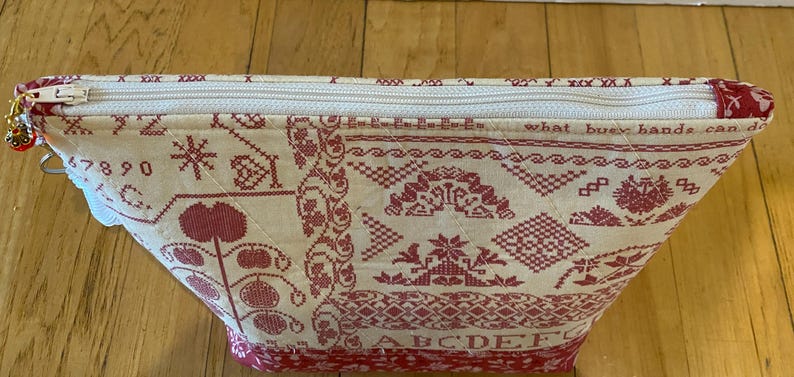 May include: A cream-colored, quilted cosmetic bag with a red cross-stitch design. The bag features a zipper closure and a decorative charm. The design includes floral and geometric patterns, along with the text "what busy hands can do."