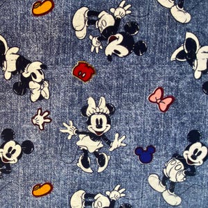 May include: A blue denim fabric featuring a repeating pattern of Mickey Mouse and Minnie Mouse characters in black and white, interspersed with small colorful icons including yellow shoes, red shorts, pink bows, and blue mouse ear silhouettes.