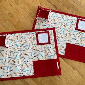 May include: Two quilted placemats with a red border and accents. The placemats feature a cream-colored background with a pattern of rolling pins in red and blue. Each has a red pocket and a small red tray.