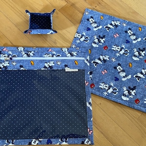 May include: A set of three fabric items featuring a blue denim-look print with Mickey and Minnie Mouse characters. The set includes a small square tray, a square quilted mat, and a square zippered pouch with a clear window and a Stitch in Peace label.