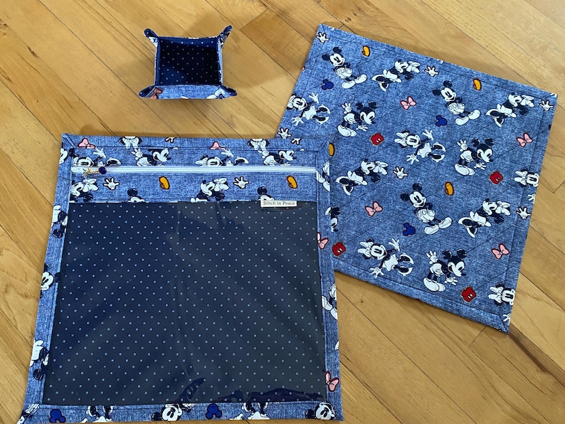 May include: A set of three fabric items featuring a blue denim-look print with Mickey and Minnie Mouse characters. The set includes a small square tray, a square quilted mat, and a square zippered pouch with a clear window and a Stitch in Peace label.