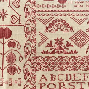 May include: Close-up of a cream-colored fabric with intricate red cross-stitch embroidery. The design includes floral patterns, geometric shapes, and the alphabet. The text "I'll show to others what busy hands" is also visible.