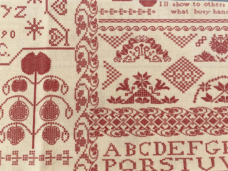 May include: Close-up of a cream-colored fabric with intricate red cross-stitch embroidery. The design includes floral patterns, geometric shapes, and the alphabet. The text "I'll show to others what busy hands" is also visible.