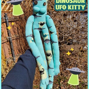 May include: A handmade teal plush toy cat with long limbs, button eyes, and a muzzle. The cat is made from socks patterned with dinosaurs and UFOs. The text "DINOSAUR UFO KITTY" is displayed in a green box.