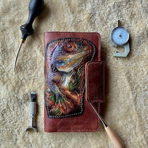 May include: A brown leather notebook cover featuring a hand-tooled, multi-coloured iguana design on the front. The cover includes a magnetic closure tab and is surrounded by leatherworking tools, including a metal thickness gauge.