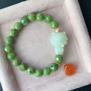 May include: A green jade bracelet with a green jade charm in the shape of a cartoon animal and a peach-colored charm. The bracelet is on a pink velvet background.