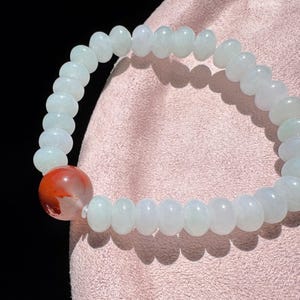 May include: A bracelet made of pale green and white rounded beads, with a single red and white bead. The bracelet is displayed on a pink surface, with a black background. The beads are approximately 0.8 cm in diameter.