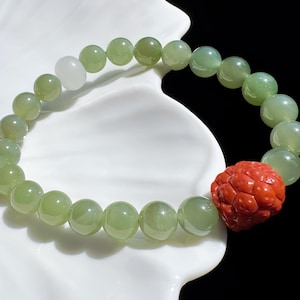 Peut inclure: Bracelet de perles rondes vertes claires et blanches. Un élément décoratif rouge sculpté est intégré. Le bracelet est présenté sur une surface blanche en forme de pétale, sur fond noir.
