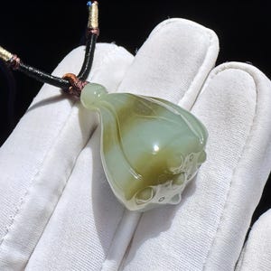May include: A jade pendant in the shape of a lotus bud, with a green and white color gradient. The pendant is suspended from a black cord with brown and gold accents. The pendant is held in a white gloved hand.