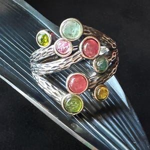 Puede incluir: Un anillo de plata con una banda texturizada y múltiples piedras redondas en tonos verdes, rosas y amarillos.