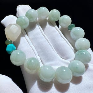 May include: A beaded bracelet featuring large, round, pale green beads. The bracelet includes a mix of other beads in shades of green, turquoise, and a faceted, off-white stone. The bracelet is displayed on a white glove.