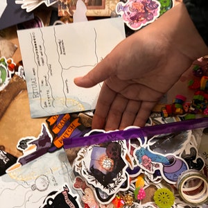 Könnte beinhalten: Nahaufnahme einer Vielzahl von Aufklebern und Bastelutensilien, darunter ein violettes Lineal, eine durchsichtige Plastiktüte gefüllt mit Aufklebern und ein Blatt Papier mit dem Wort "RITUAL" darauf. Die Aufkleber zeigen verschiedene Themen, darunter Hexen, Monde und Blumen.