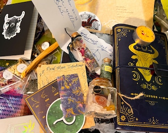Op de afbeelding: Een blauw en gouden dagboek met een stierenontwerp en het woord "Taurus" op de omslag. Het dagboek wordt omringd door verschillende items, waaronder een klein flesje gedroogde bloemen, een klein potje groene kristallen, een klein potje kristallen, een houten stok en een stuk papier met de woorden "27 New Moon Intentions".