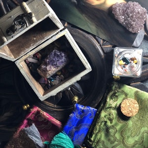 Pode incluir: Uma caixa de madeira cheia de vários cristais e pedras, incluindo uma grande ametista, está sobre uma superfície escura.  Três pequenas bolsas com borlas também estão sobre a superfície, uma é verde, outra azul e outra bordô.  Um pequeno frasco de vidro com tampa de cortiça contém uma variedade de cristais e pedras.  Uma impressão emoldurada de uma mulher com uma coroa de lua crescente está no fundo.