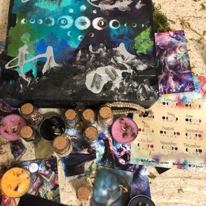Könnte beinhalten: Eine Holzkiste mit einem schwarzen und blauen abstrakten Gemälde auf dem Deckel. Die Kiste ist geöffnet und enthält kleine Glasflaschen mit Korkverschlüssen, Kerzen und Tarotkarten. Die Karten zeigen Bilder des Mondes und anderer Himmelskörper. Die Kiste enthält auch einen Kalender mit den Mondphasen für jeden Monat des Jahres.