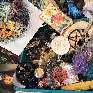 May include: A teal box filled with various items, including a hand-painted wheel of the year, a wooden circle with a pentagram, a small bottle with a cork stopper, a tarot card, and a variety of crystals and stones.