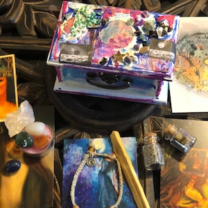 Op de afbeelding: Een klein houten doosje met een kleurrijk geschilderd ontwerp, met een maan en sterren. Het doosje is versierd met kristallen en andere versieringen. Het doosje is omringd door verschillende voorwerpen, waaronder een klein flesje kruiden, een houten stok, een armband en een kaart met een maan- en sterrenmotief.