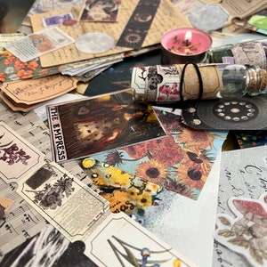 Puede incluir: Una colecci&oacute;n de ef&iacute;meras vintage, que incluye cartas del tarot, etiquetas bot&aacute;nicas, partituras y cinta decorativa. Tambi&eacute;n hay una vela rosa encendida y peque&ntilde;os frascos de vidrio con tapones de corcho. Los art&iacute;culos est&aacute;n dispuestos sobre una superficie.