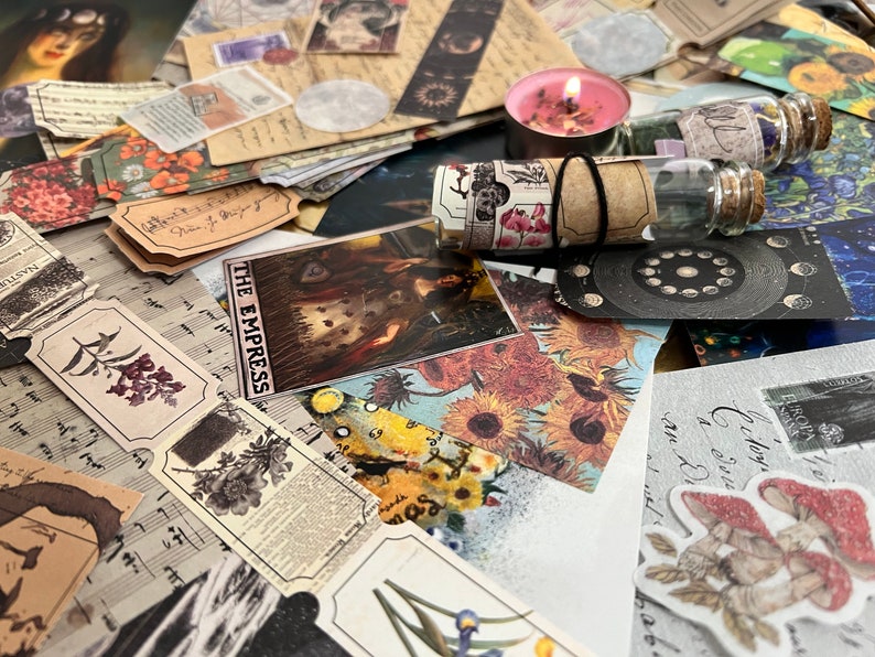 Puede incluir: Una colecci&oacute;n de ef&iacute;meras vintage, que incluye cartas del tarot, etiquetas bot&aacute;nicas, partituras y cinta decorativa. Tambi&eacute;n hay una vela rosa encendida y peque&ntilde;os frascos de vidrio con tapones de corcho. Los art&iacute;culos est&aacute;n dispuestos sobre una superficie.