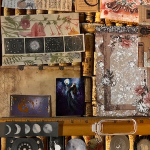 Puede incluir: Una colecci&oacute;n de ef&iacute;meras decorativas de papel con temas florales, celestiales y vintage. Incluye estampados bot&aacute;nicos, ilustraciones de fases lunares y una peque&ntilde;a pintura de una figura sosteniendo una luna. La paleta de colores presenta tonos apagados con toques de color.