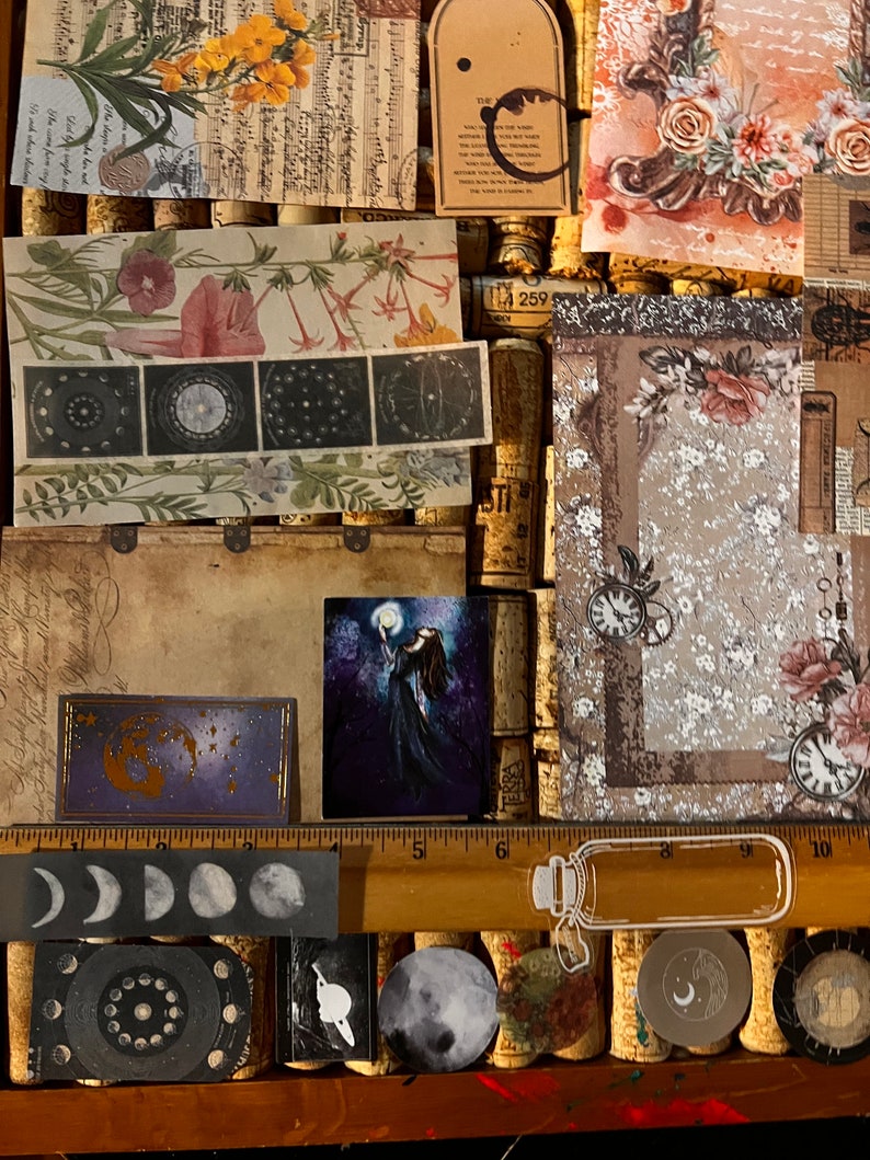 Puede incluir: Una colecci&oacute;n de ef&iacute;meras decorativas de papel con temas florales, celestiales y vintage. Incluye estampados bot&aacute;nicos, ilustraciones de fases lunares y una peque&ntilde;a pintura de una figura sosteniendo una luna. La paleta de colores presenta tonos apagados con toques de color.
