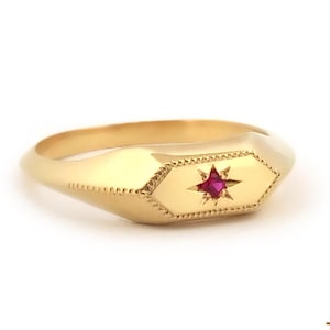 May include: A gold ring with a ruby-colored starburst design set in an octagonal shape. The ring has a smooth, polished band and a detailed, textured design around the starburst. The ring is displayed on a white background.
