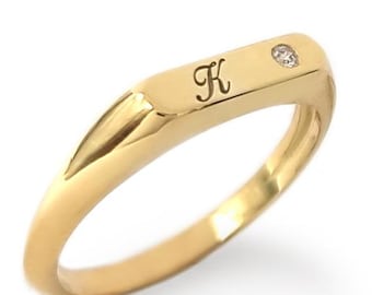 May include: A gold signet ring featuring a flat top surface engraved with the letter K in a cursive script font. A small, round, clear gemstone is set into the metal surface next to the letter. The band has a smooth, polished finish.