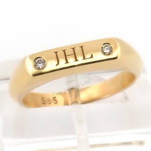 Könnte beinhalten: Ein goldener Ring mit den Initialen "JHL" auf der Oberseite eingraviert, flankiert von zwei kleinen Diamanten. Der Ring hat eine polierte Oberfläche und wird auf einem transparenten Ständer präsentiert. Der Ring ist im Siegelring-Stil.