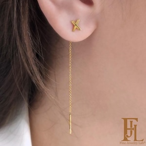 Op de afbeelding: Een paar goudkleurige oorbellen. Eén is een X-vormige oorbel, de andere een lange ketting met een kleine staaf. De oorbellen worden op het oor van een persoon gedragen.