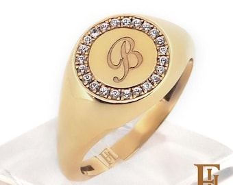 May include: A gold signet ring with a round face. The face features a capital letter "B" in a script font, surrounded by a circle of small, clear gemstones. The ring is displayed on a white surface.