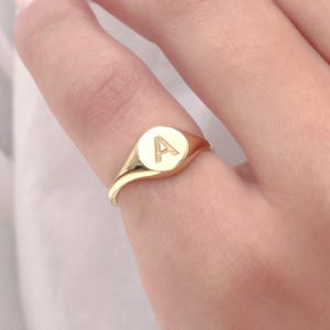 May include: A gold signet ring with the letter "A" engraved on a circular face. The ring has a polished finish and is worn on a finger. The ring is a classic piece of jewelry.
