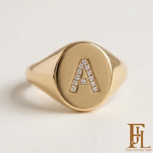Könnte beinhalten: Ein goldener Siegelring mit ovaler Front und einem mittig platzierten Buchstaben A. Der Buchstabe A ist mit kleinen, klaren, funkelnden Steinen besetzt. Die Ringschiene und die Vorderseite haben eine glatte, polierte Goldoptik.