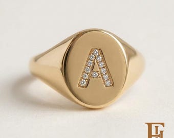 May include: A gold signet ring featuring an oval face with a centered letter A. The letter A is inlaid with small, clear, sparkling stones. The band and the face of the ring have a smooth, polished, reflective gold-toned metal finish.