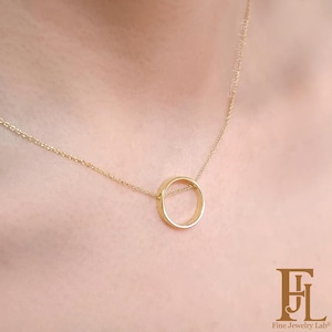 Puede incluir: Un delicado collar de oro con un colgante circular. El colgante es un diseño de círculo abierto simple, suspendido de una fina cadena. El collar se muestra sobre un fondo color piel, resaltando la elegante simplicidad de la joya.