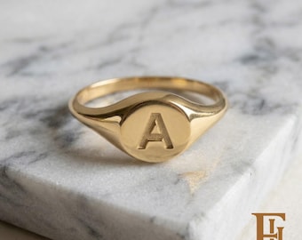 Op de afbeelding: Een gepolijste gouden zegelring met een cirkelvormig vlak waarin de hoofdletter A is gegraveerd. De ring heeft een gladde, reflecterende metalen afwerking en een taps toelopend bandontwerp voor een strak en eenvoudig uiterlijk.