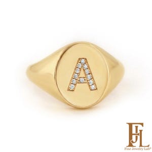 Peut inclure: Bague chevalière en or avec la lettre "A" incrustée de petites pierres scintillantes. La bague a une face ovale polie et un anneau lisse et arrondi. Un bijou classique.