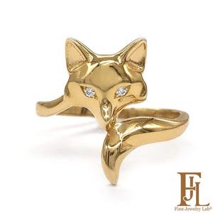 Può includere: Un anello color oro a forma di testa di volpe con due piccole pietre rotonde e chiare come occhi. Il design della volpe presenta orecchie a punta e una coda curva che forma la fascia dell'anello. Il testo Fine Jewelry Lab è visibile.