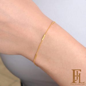 May include: A delicate gold bracelet featuring a fine chain with three rectangular bar accents. The bracelet is worn on a wrist, showcasing its minimalist design and subtle elegance. Fine Jewelry Lab logo is visible.
