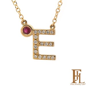 Può includere: Collana color oro con un ciondolo a forma di lettera «E». La lettera è incastonata con piccoli diamanti scintillanti. Una piccola gemma rossa rotonda è posta a sinistra della lettera. La collana è su sfondo bianco.