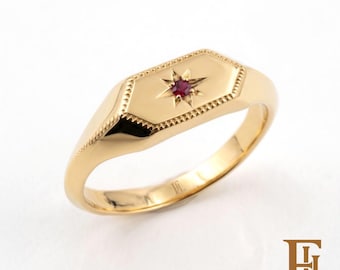 May include: A gold signet ring featuring a hexagonal face with a beaded border. A small, round, deep red gemstone is set in the centre, surrounded by an engraved eight-pointed starburst design. The interior of the band is stamped with the letters FL.