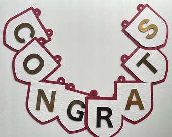 May include: A banner spelling "CONGRATS" in gold and black letters. Each letter is on a white, textured pennant with a burgundy border. The pennants are connected with small burgundy loops at the top, creating a celebratory garland.