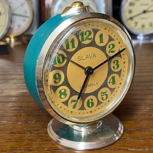 May include: A vintage teal and silver alarm clock with a gold-coloured face. The clock face features green numerals and the brand name "SLAVA". The clock has a round shape and a silver base.