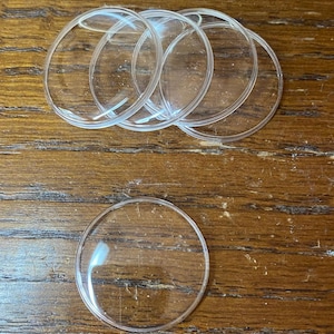 May include: A collection of five clear, circular, convex glass or plastic cabochons. These transparent, dome-shaped discs feature smooth, rounded edges and a polished surface, designed for crafting or decorative applications in various projects.