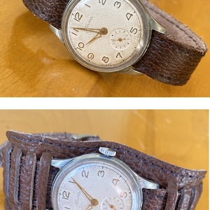 May include: A vintage wristwatch with a silver case and a white face, displaying black numerals and hands. It features a small seconds dial and is secured by a wide, brown leather cuff strap. The watch face reads "Kama".