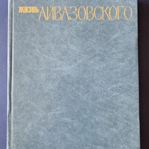 Op de afbeelding: Een hardcover boek met een getextureerde, grijsblauwe kaft. De titel, in gouden letters, luidt "ЖИЗНЬ АЙВАЗОВСКОГО". De rug van het boek is aan de linkerkant zichtbaar. De kaft heeft een licht versleten uiterlijk.