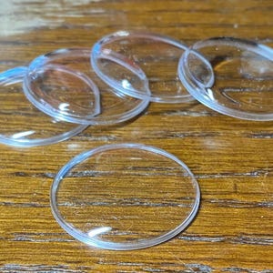 May include: A collection of five clear, circular, dome-shaped glass cabochons. Each piece features a smooth, transparent surface with a rounded top and a flat base, designed for crafting or decorative applications, arranged in a loose cluster.