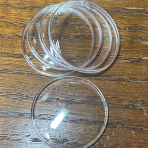May include: A collection of five circular, transparent, and slightly domed glass or plastic watch crystals. These clear, round components feature smooth, polished edges and a reflective surface, designed as replacement parts for timepieces.