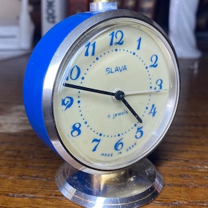 Könnte beinhalten: Ein blauer und silberner Vintage-Wecker mit cremefarbenem Zifferblatt. Das Zifferblatt hat blaue Ziffern, das Wort "SLAVA" und den Text "11 jewels MADE IN USSR". Der Wecker hat einen silbernen Sockel und eine goldfarbene Oberseite.