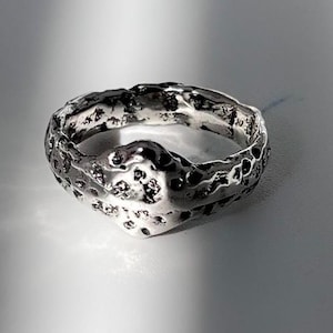 Peut inclure: Une bague argentée avec une surface très texturée et irrégulière, présentant des creux profonds et des bosses organiques en relief. Le métal possède une finition polie et réfléchissante qui capte la lumière, créant un contraste marqué entre les crevasses sombres et les reflets métalliques brillants sur l'anneau.