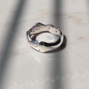 May include: A silver ring with an irregular, wavy, and organic band shape. The metal surface is polished and reflective, showing subtle indentations and a smooth, flowing contour that creates a non-uniform, undulating profile around the finger opening.